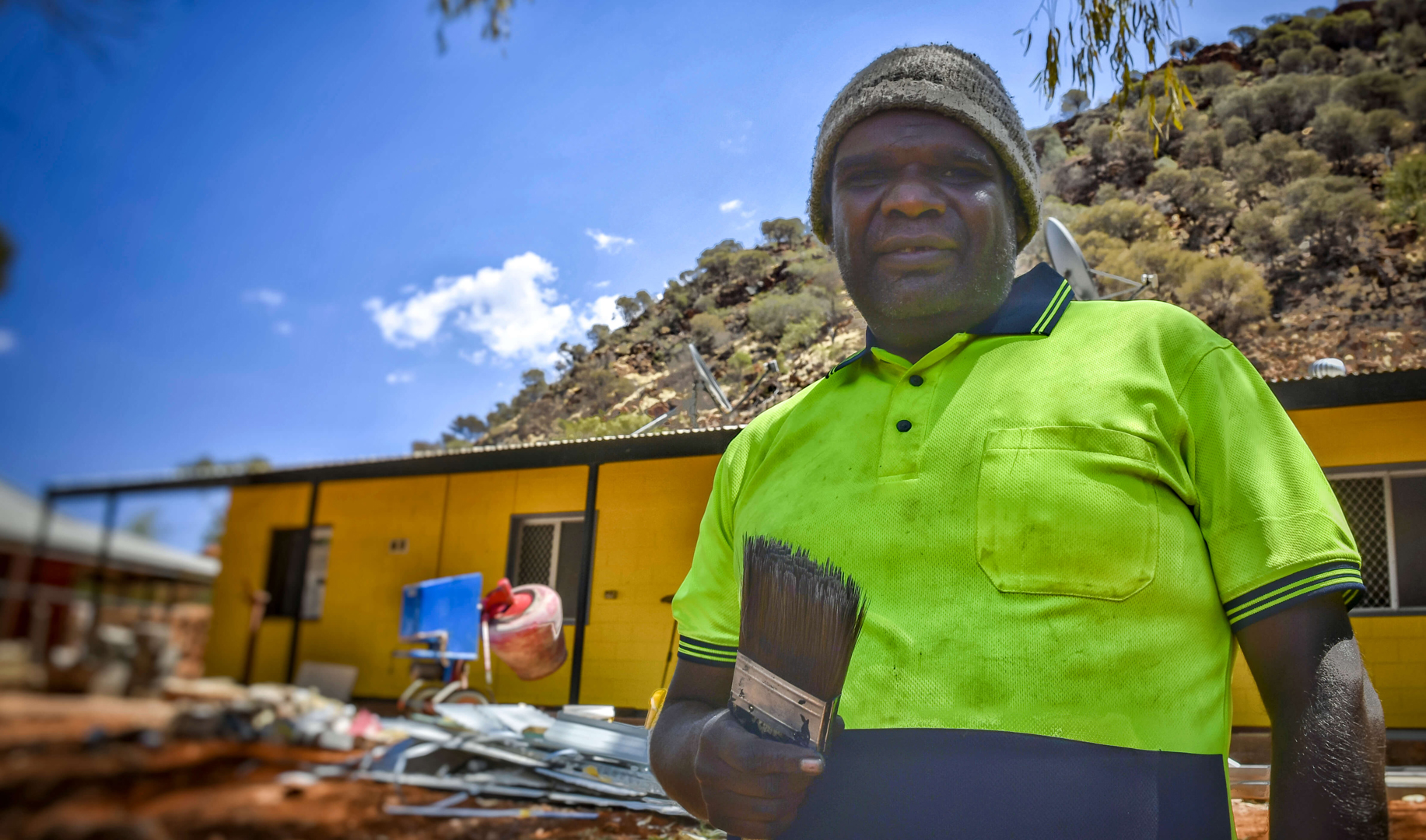 Indigenous man holding paintbrush in front of local house.